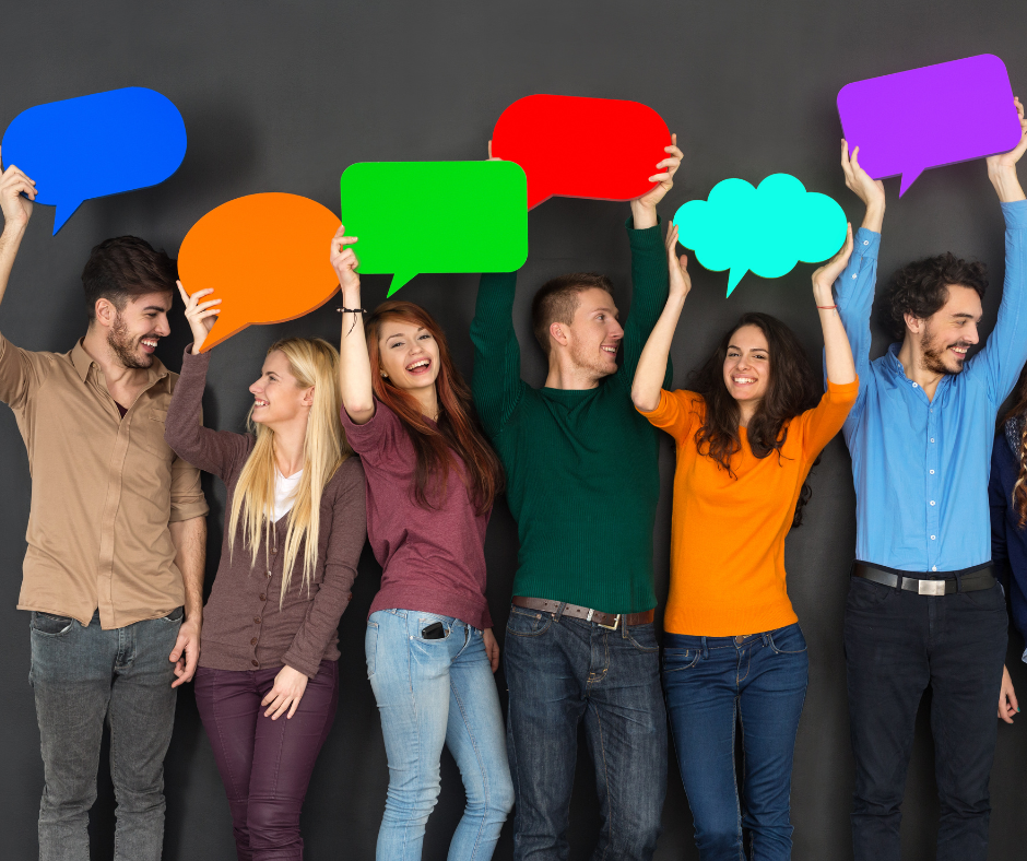Zdjęcie przedstawia Grupę młodych, uśmiechniętych ludzi stojących obok siebie na tle ciemnej ściany, trzymających nad głowami kolorowe dymki komiksowe, symbolizujące wypowiedzi lub komunikację.