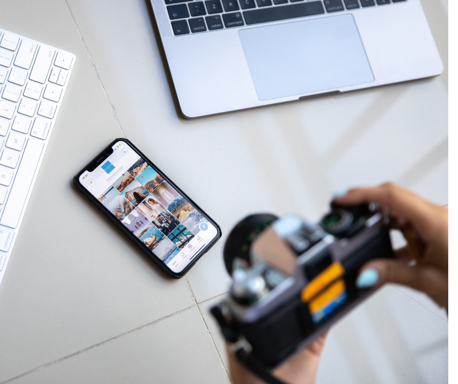 Zdjęcie przedstawia aparat fotograficzny, smartphone oraz laptopa na biurku.