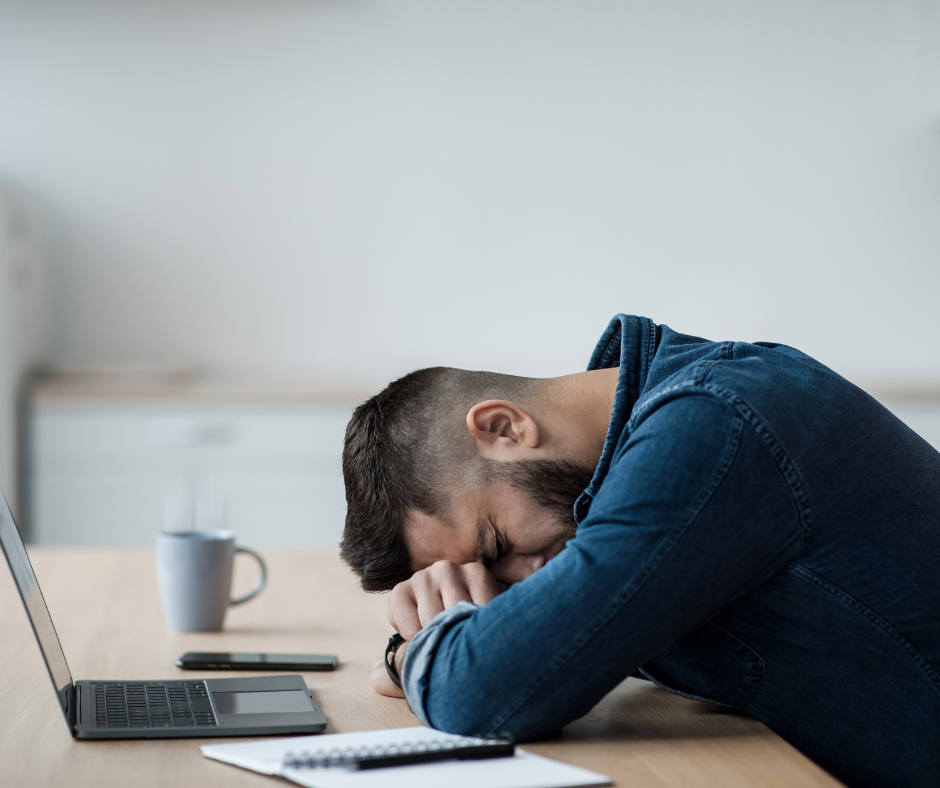 Zdjęcie przedstawia wyczerpanego mężczyznę przy komputerze opierającego głowę o biurko.