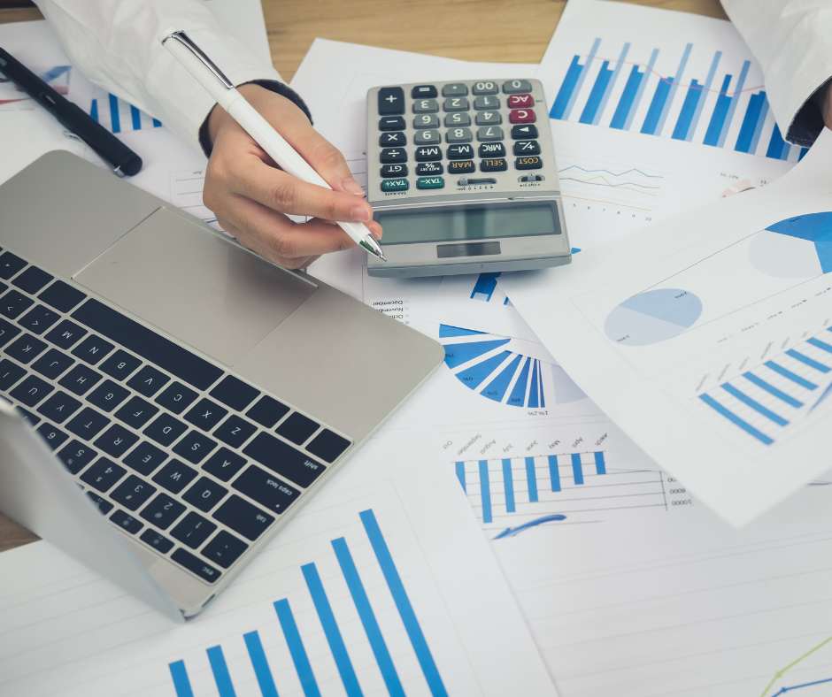 Zdjęcie przedstawia rękę trzymającą długopis i kalkulator na biurku pełnym dokumentów biznesowych.