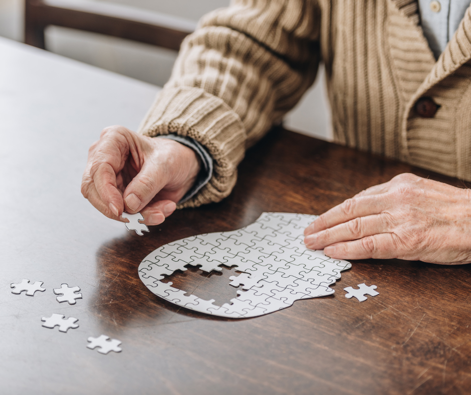 Zdjęcie przedstawia zbliżenie na dłonie starszego mężczyzny, który układa białe puzzle w kształcie głowy.