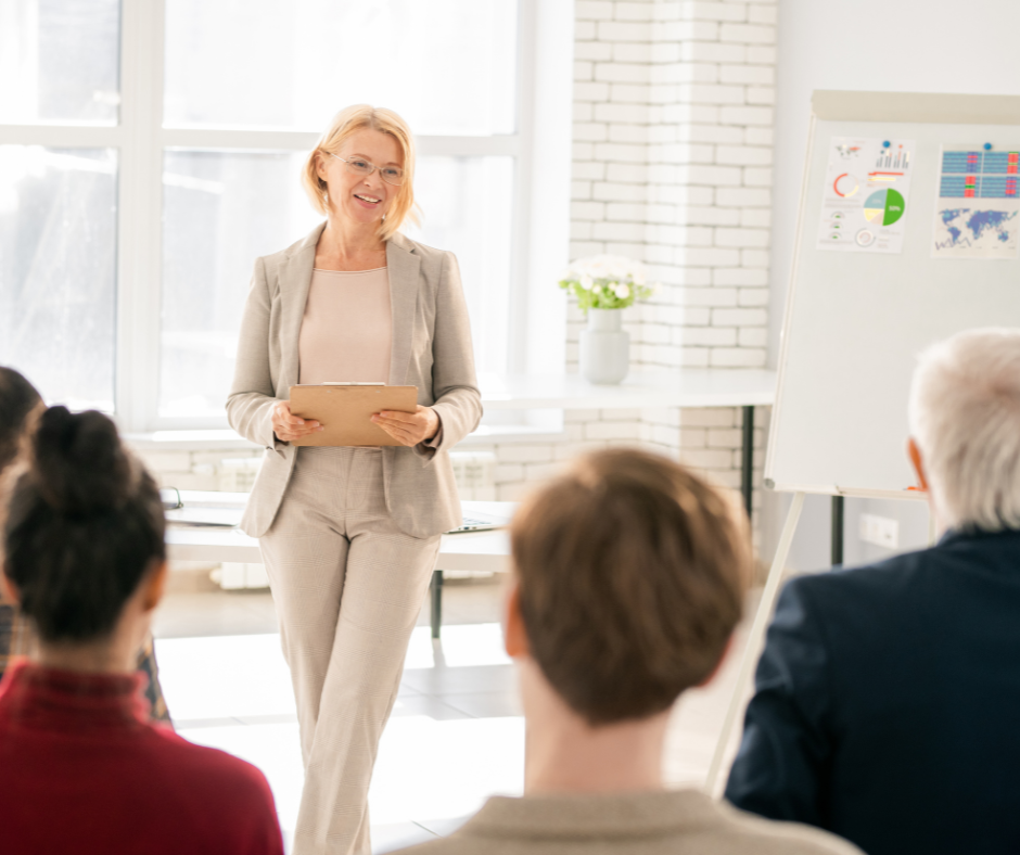 Zdjęcie przedstawia formalnie ubraną w jasne kolory kobietę prowadzącą szkolenie dla grupy ludzi. W tle notatki.