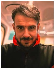 Zdjęcie przedstawia mężczyznę z krótkimi brązowymi włosami i zarostem, w czarnej kurtce.