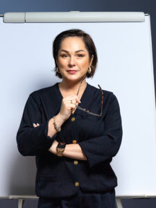 Zdjęcie przedstawia kobietę z krótkimi włosami, ubraną w czarną koszulę. W tle tablica szkoleniowa.