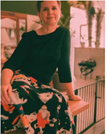 Kobieta z krótkimi włosami, ubrana w ciemną bluzkę z rękawem 3/4 i kolorową spódnicę z graficznymi postaciami, siedzi na drewnianym blacie w jasnym, nowoczesnym wnętrzu z roślinami w tle.