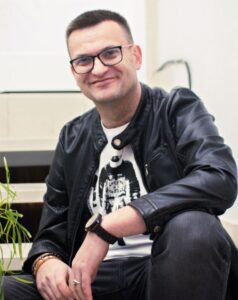 Zdjęcie przedstawia Mężczyznę w okularach i czarnej skórzanej kurtce, siedzi ze swobodnym uśmiechem. Ma na sobie koszulkę z grafiką astronauty i ciemne spodnie. W tle jasne wnętrze i roślina po lewej stronie kadru