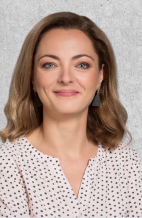 Zdjęcie przedstawia dorosłą kobietę z blond włosami do ramion, uśmiechnięta, ubrana w białą koszulę w kropki.