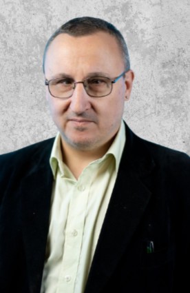 Zdjęcie przedstawia mężczyznę w średnim wieku z bardzo krótkimi, ciemnymi włosami i okularami. Ma na sobie garnitur.