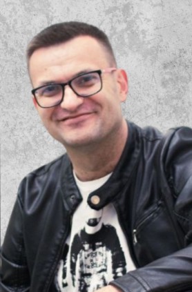 Zdjęcie przedstawia mężczyznę z krótkimi włosami w okularach. Jest uśmiechnięty i ma na sobie skórzaną kurtkę i białą koszulkę.