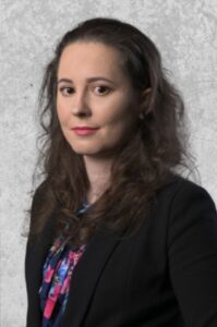 Zdjęcie przedstawia uśmiechniętą dorosłą kobietę z długimi lekko kręconymi, ciemnymi włosami. Jest ubrana w czarną marynarkę i wzorzystą koszulę.