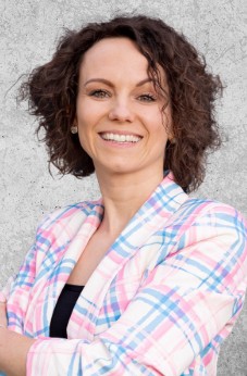 Zdjęcie przedstawia uśmiechniętą kobietę z kręconymi włosami do ramion, ubrana we wzorzystą marynarkę.