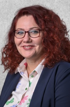 Zdjęcie przedstawia uśmiechniętą kobietę w okularach o kręconych rudych włosach. Ubrana w koszulę z kwiatowym wzorem i marynarkę.