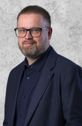Zdjęcie przedstawia uśmiechniętego mężczyznę w okularach, krótkimi włosami i brodą. Ubrany formalnie w koszulę i marynarkę.