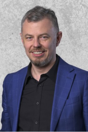 Zdjęcie przedstawia mężczyznę z krótkimi siwymi włosami i brodą. Ubrany w czarną koszulę i granatową marynarkę. Uśmiechnięty.