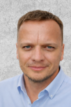 Zdjęcie przedstawia mężczyznę o krótkich blond włosach i krótkim zaroście. Ubrany w niebieską koszulę.